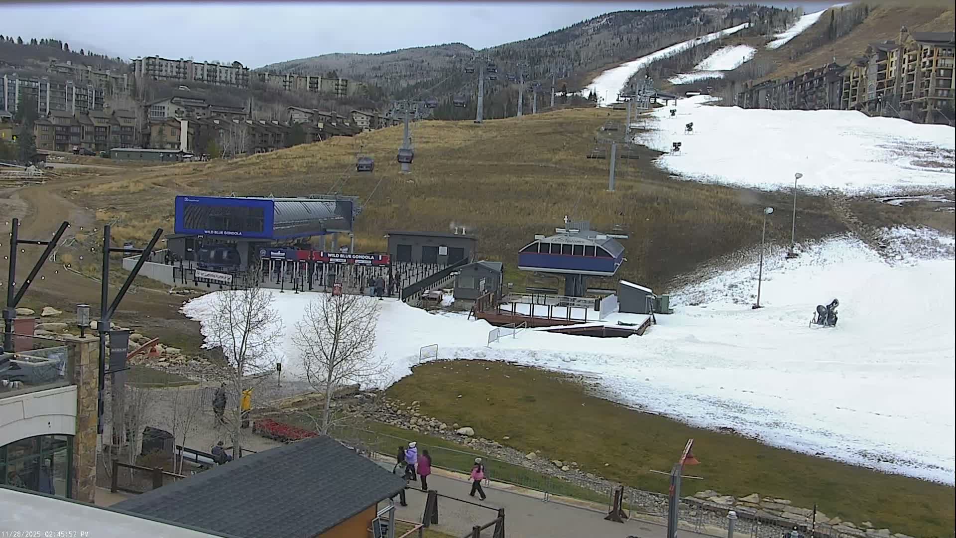 Under an overcast sky, a ski resort reveals patchy artificial snow on brown slopes with visible snowmaking machines, a gondola and chairlift station, and various resort buildings and pedestrians at the base.
