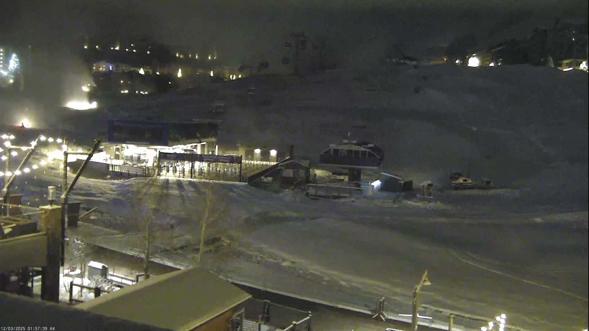 A nighttime view of a snowy ski resort reveals illuminated buildings, active ski lifts, and a snow groomer on the slopes, with mist or fog present, likely from a snow cannon in operation.