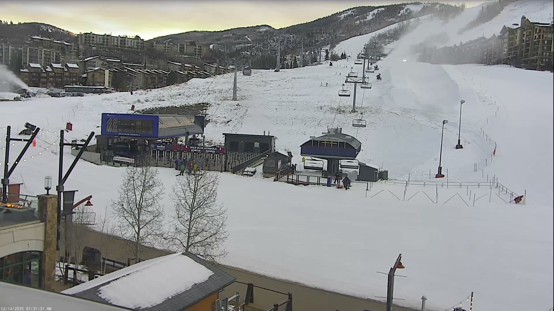 A nighttime view of a snowy ski resort reveals illuminated buildings, active ski lifts, and a snow groomer on the slopes, with mist or fog present, likely from a snow cannon in operation.