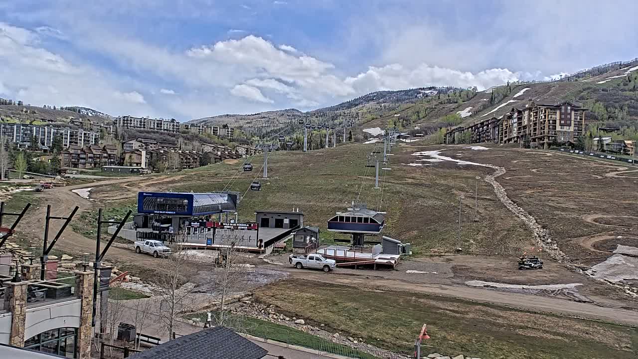 A nighttime view of a snowy ski resort reveals illuminated buildings, active ski lifts, and a snow groomer on the slopes, with mist or fog present, likely from a snow cannon in operation.