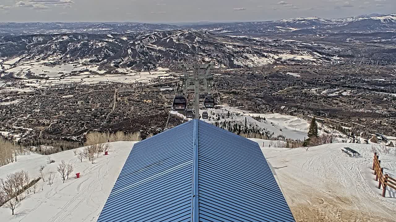 A blue-roofed structure overlooks a snowy mountain landscape with a chairlift descending toward a town below under a partly cloudy sky.