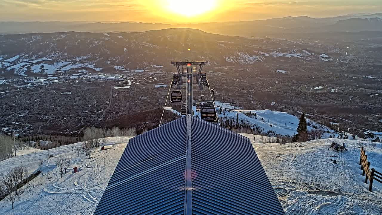 A ski lift with several gondolas ascends a snowy mountain at sunset, overlooking a valley town below.