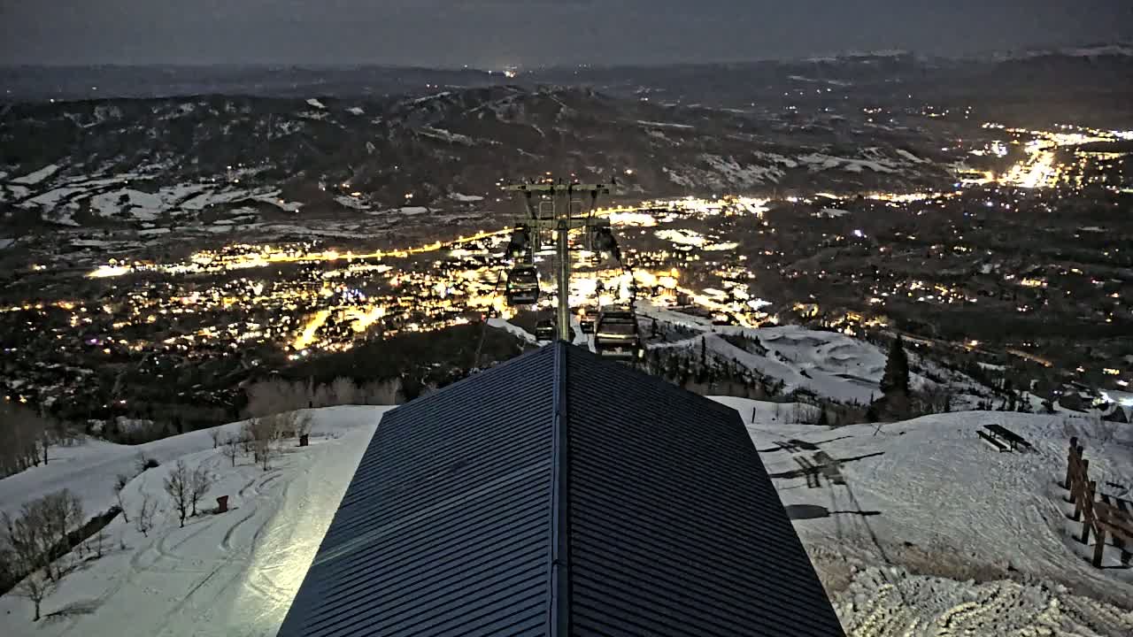 A dark, snow-covered mountain slope at night overlooks a valley town illuminated by lights, with a chairlift and a building's dark roof visible in the foreground.