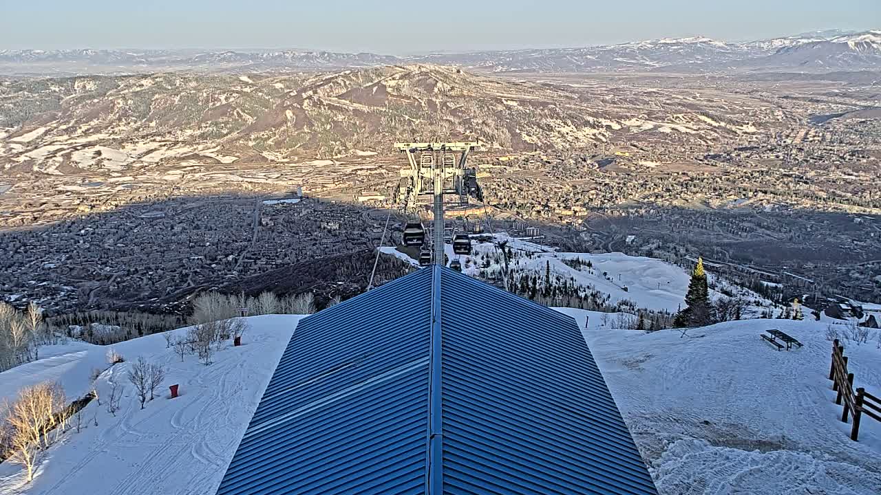 A blue roof of a building overlooks a snow-covered mountainside with a ski lift and a valley town below under a clear, sunny sky.
