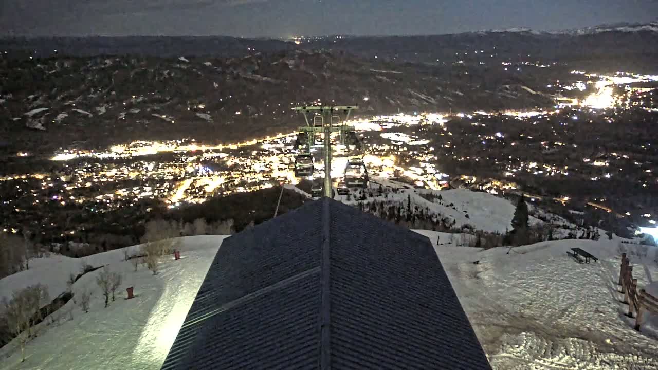 A nighttime view from a snowy mountaintop shows a chairlift overlooking a brightly lit town below.