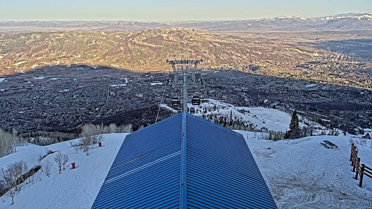 A blue-roofed structure sits atop a snowy mountain slope overlooking a valley town and distant mountains under a clear sky.