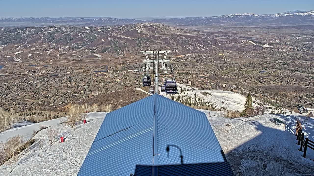 A chairlift ascends a snowy mountainside overlooking a valley town under a clear blue sky.