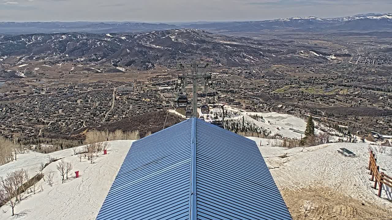A blue metal roof of a structure overlooks a snow-covered mountainside, a ski resort, and a valley town under a partly cloudy sky.