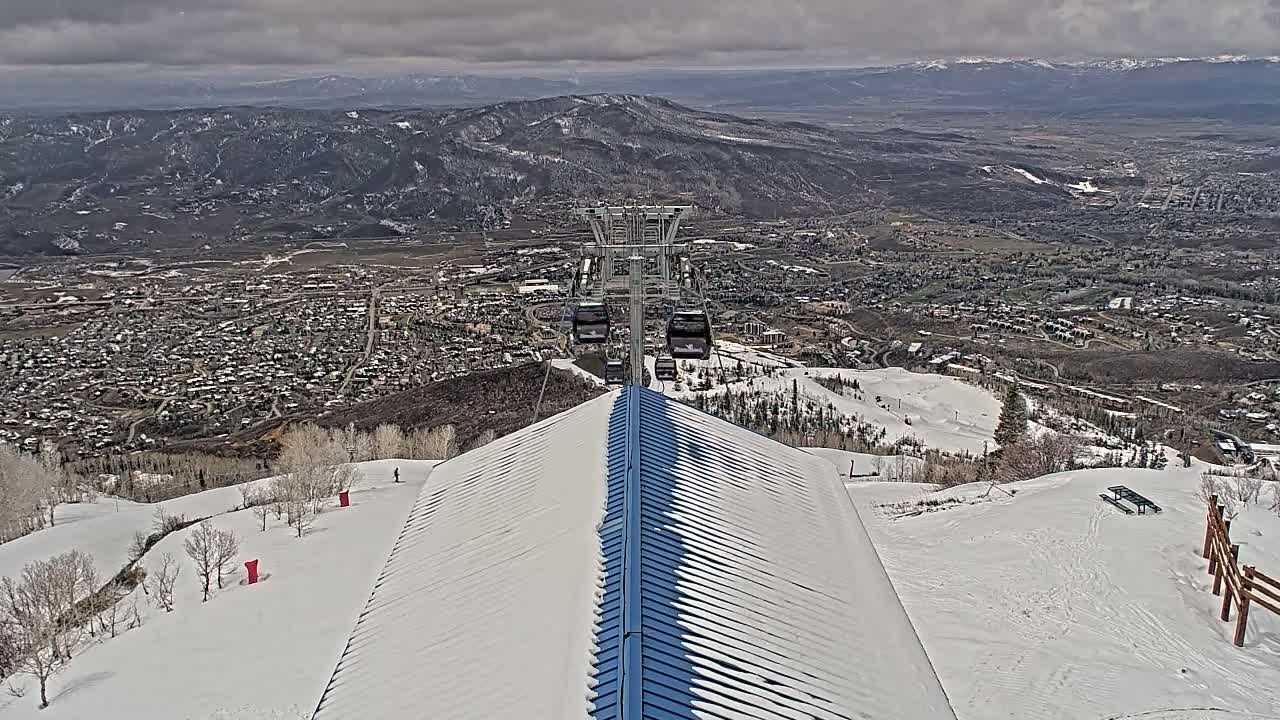 A snow-covered mountaintop with a partially snow-covered building and a ski lift ascending towards a valley town under a cloudy sky.