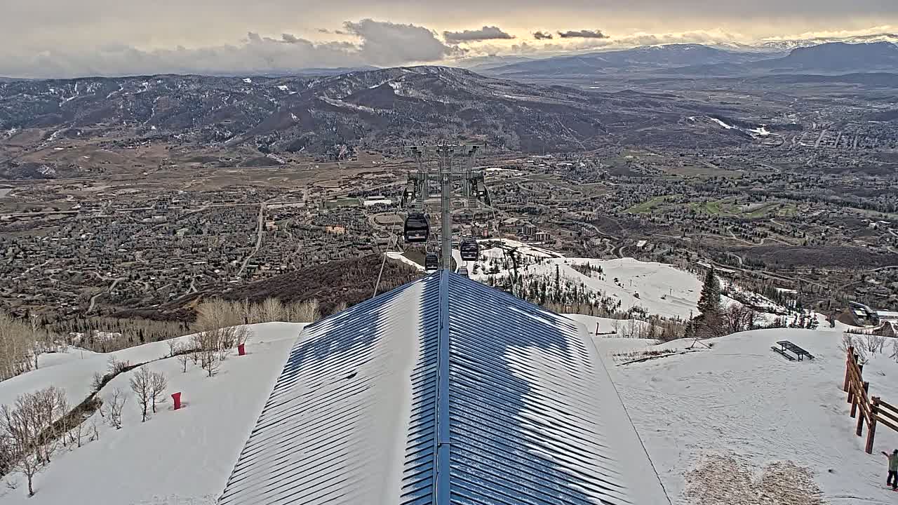 A snow-covered mountaintop with a chairlift and a view of a valley town under a cloudy sky.