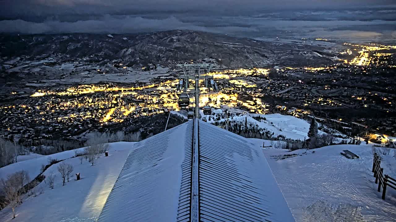 A snow-covered mountaintop building overlooks a valley town illuminated at night, with a ski lift and a dark, cloudy sky.