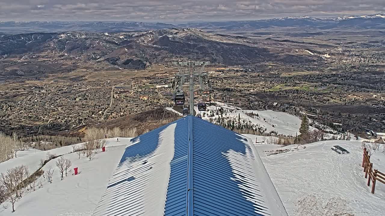 A chairlift ascends a snowy mountain overlooking a valley town under a partly cloudy sky.
