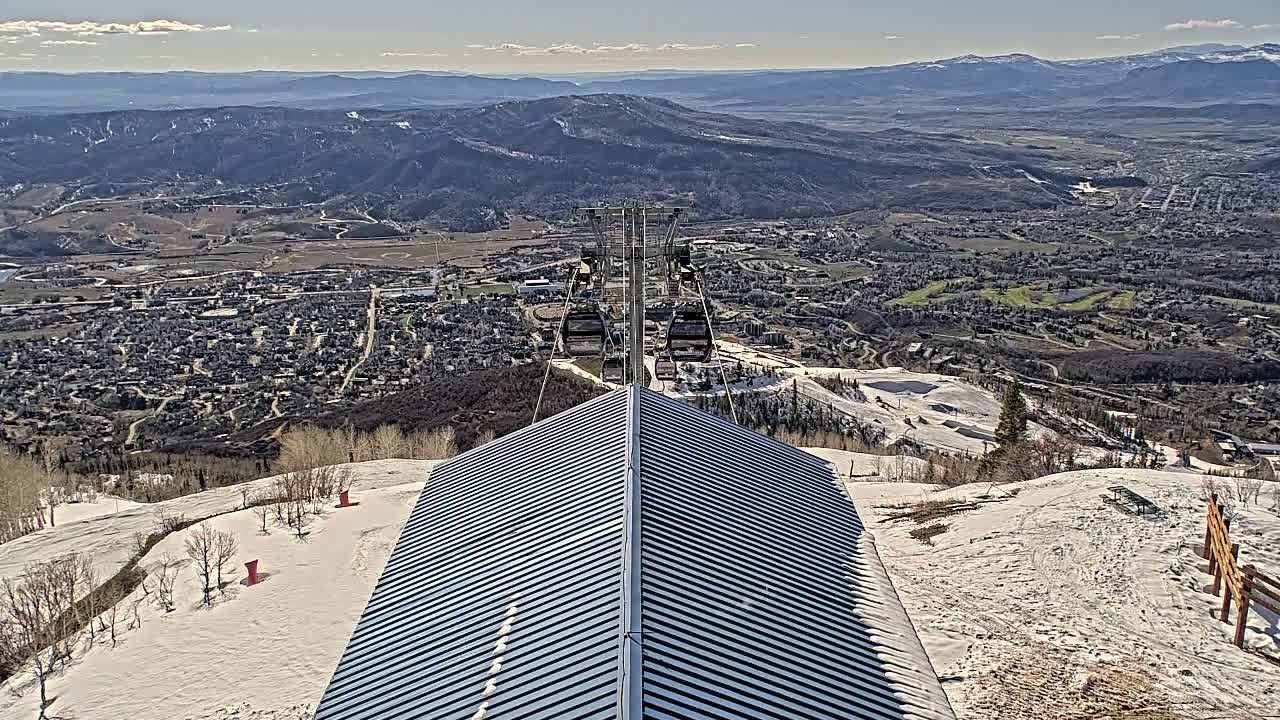 A mountaintop view on a sunny day shows a ski resort with a chairlift and a metal roofed building overlooking a valley town and distant mountains.