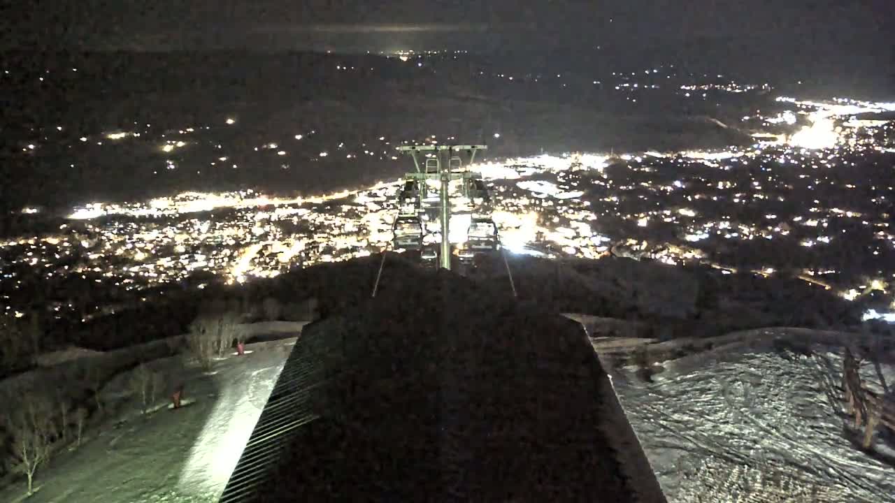 A nighttime view from a ski lift overlooking a brightly lit town below, with snow on the ground.