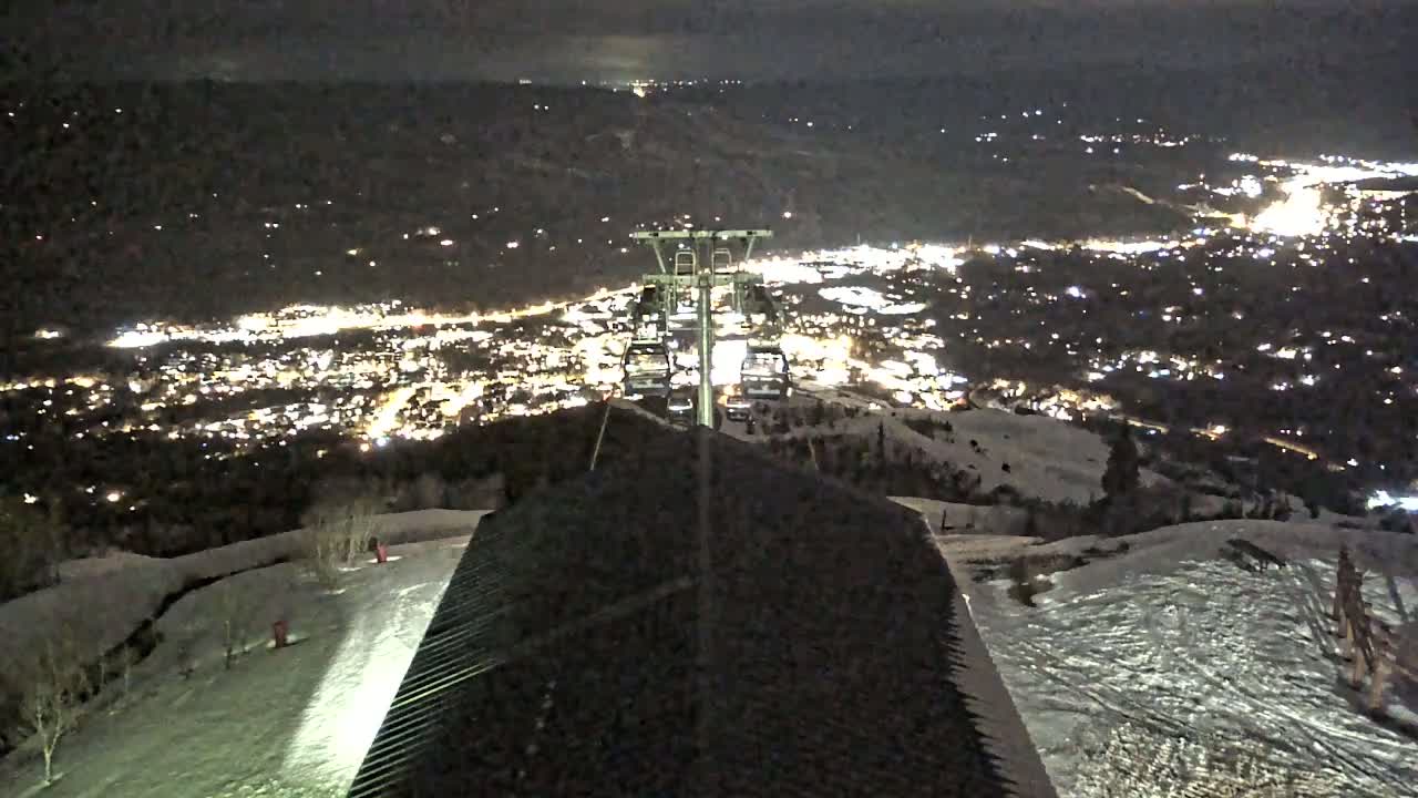 A nighttime view from a snowy mountaintop shows a chairlift ascending towards a brightly lit town in the valley below.