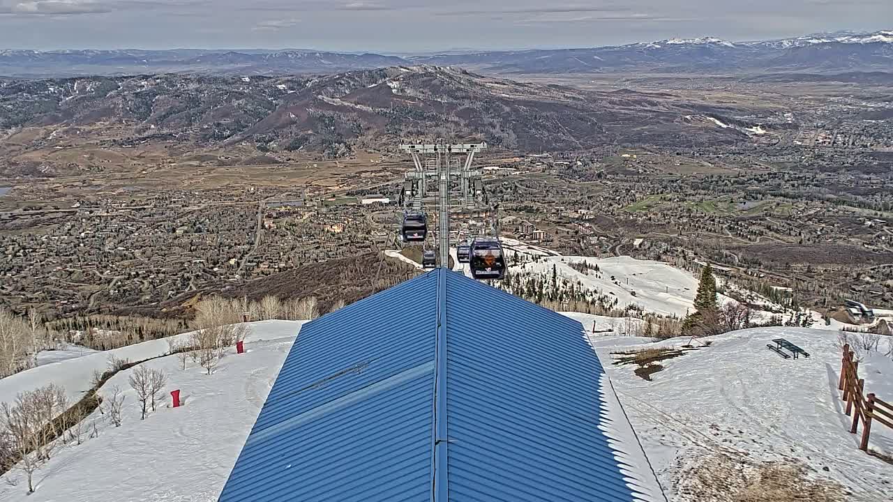 A blue roofed structure on a snow-covered mountain overlooks a valley town under a cloudy sky.