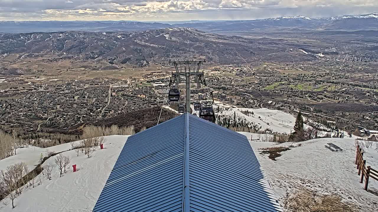 A ski lift carrying several gondolas ascends above a snow-covered mountainside overlooking a valley town under a partly cloudy sky.
