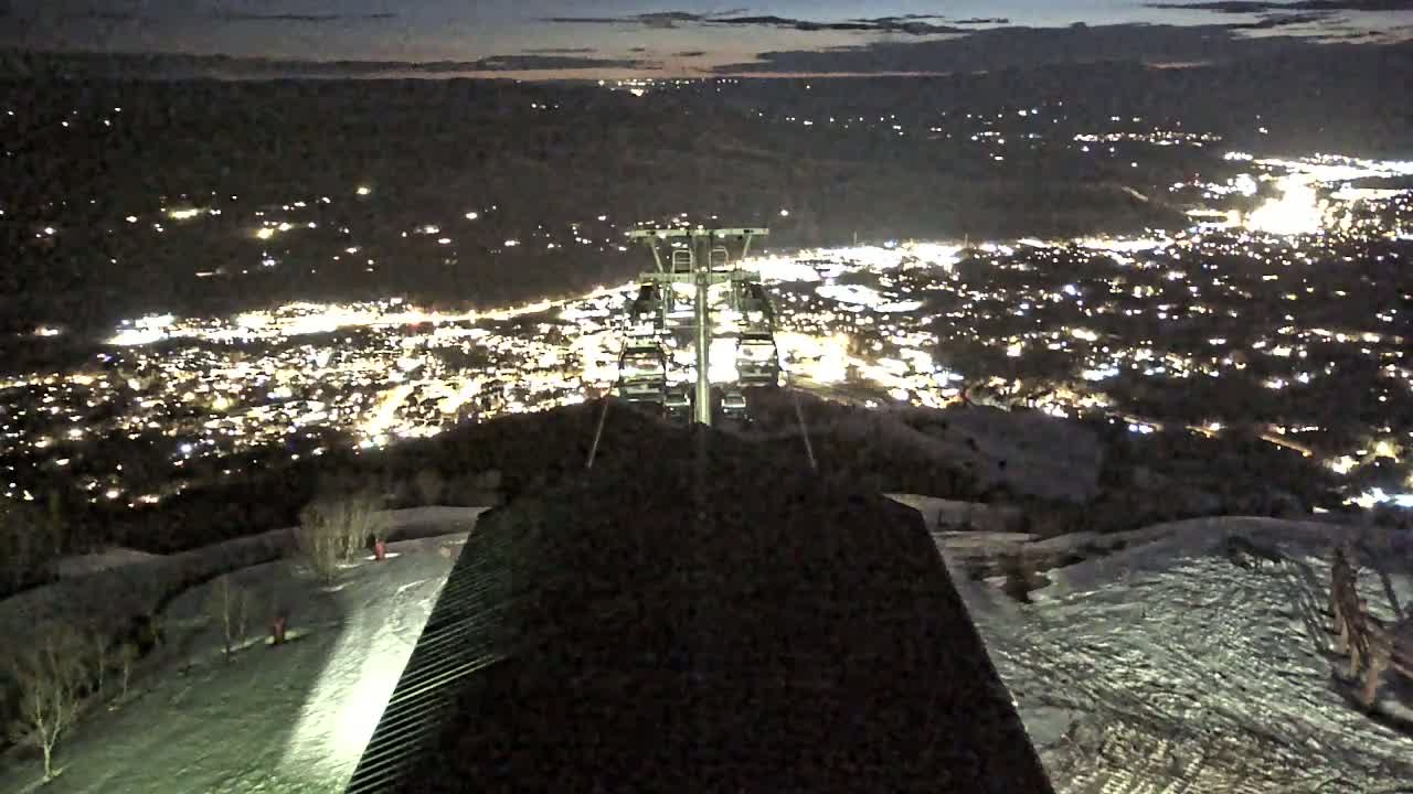 A nighttime view from a ski lift overlooking a brightly lit town below, with snow on the ground.