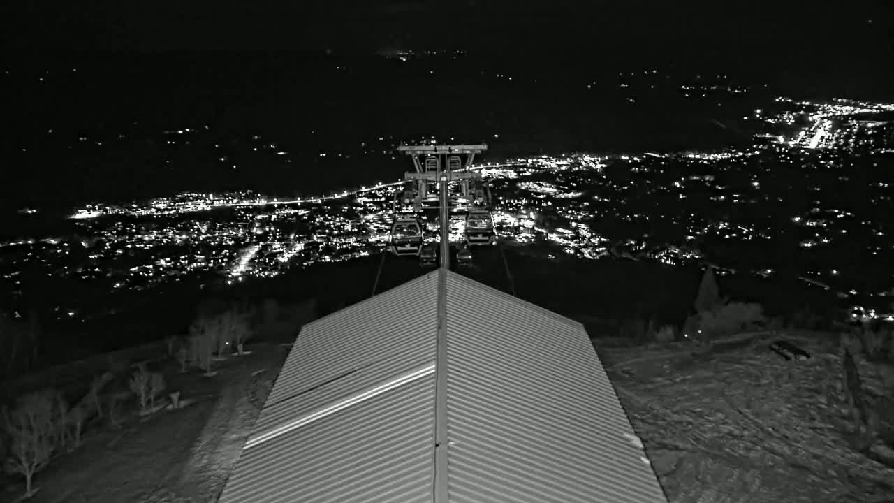 A nighttime view from a ski lift shows a city illuminated below, with snow on the ground around a building's roof.