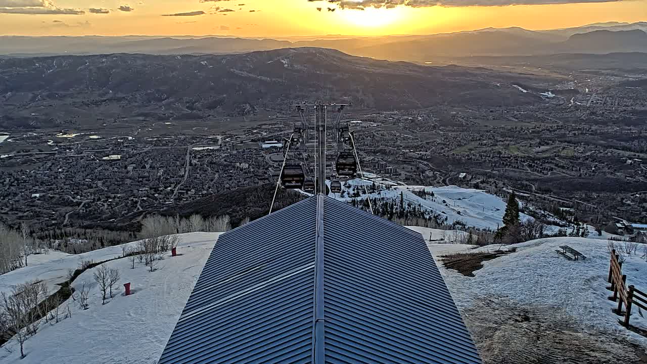 A ski lift ascends a mountain at sunset, overlooking a valley town partially covered in snow.