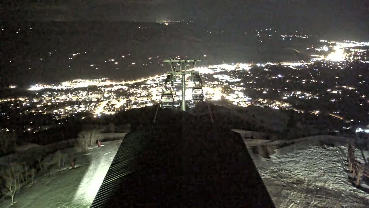 A nighttime view from a ski lift shows a snow-covered hillside and a brightly lit town below.