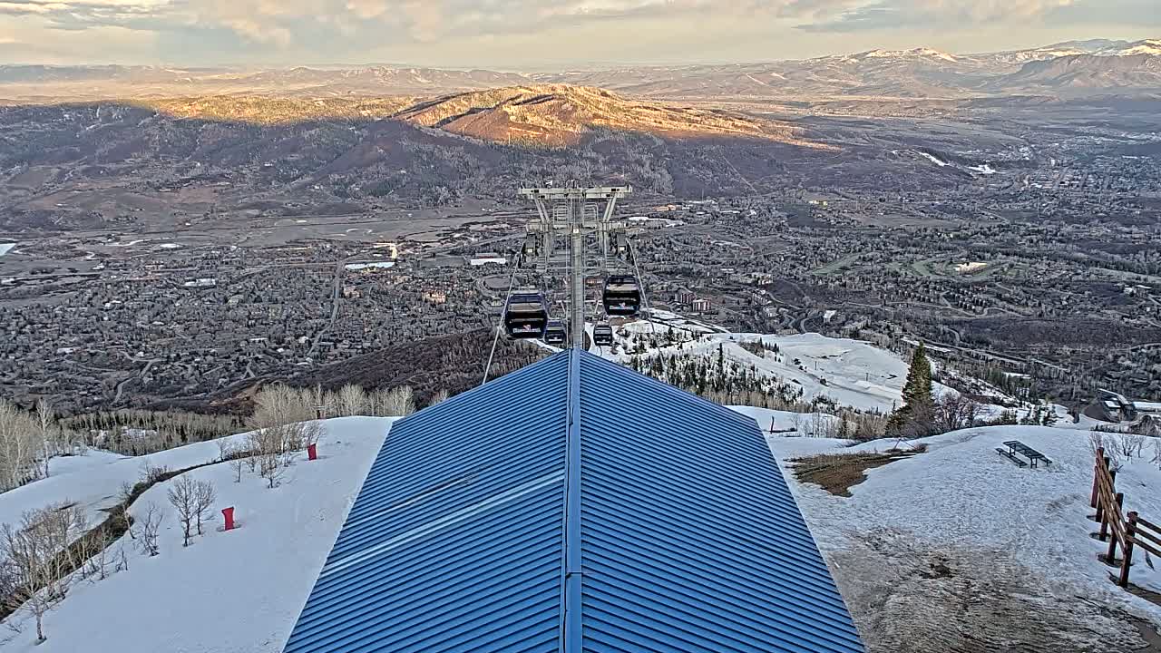 A blue-roofed building sits atop a snow-covered mountain overlooking a valley town under a partly cloudy sky.