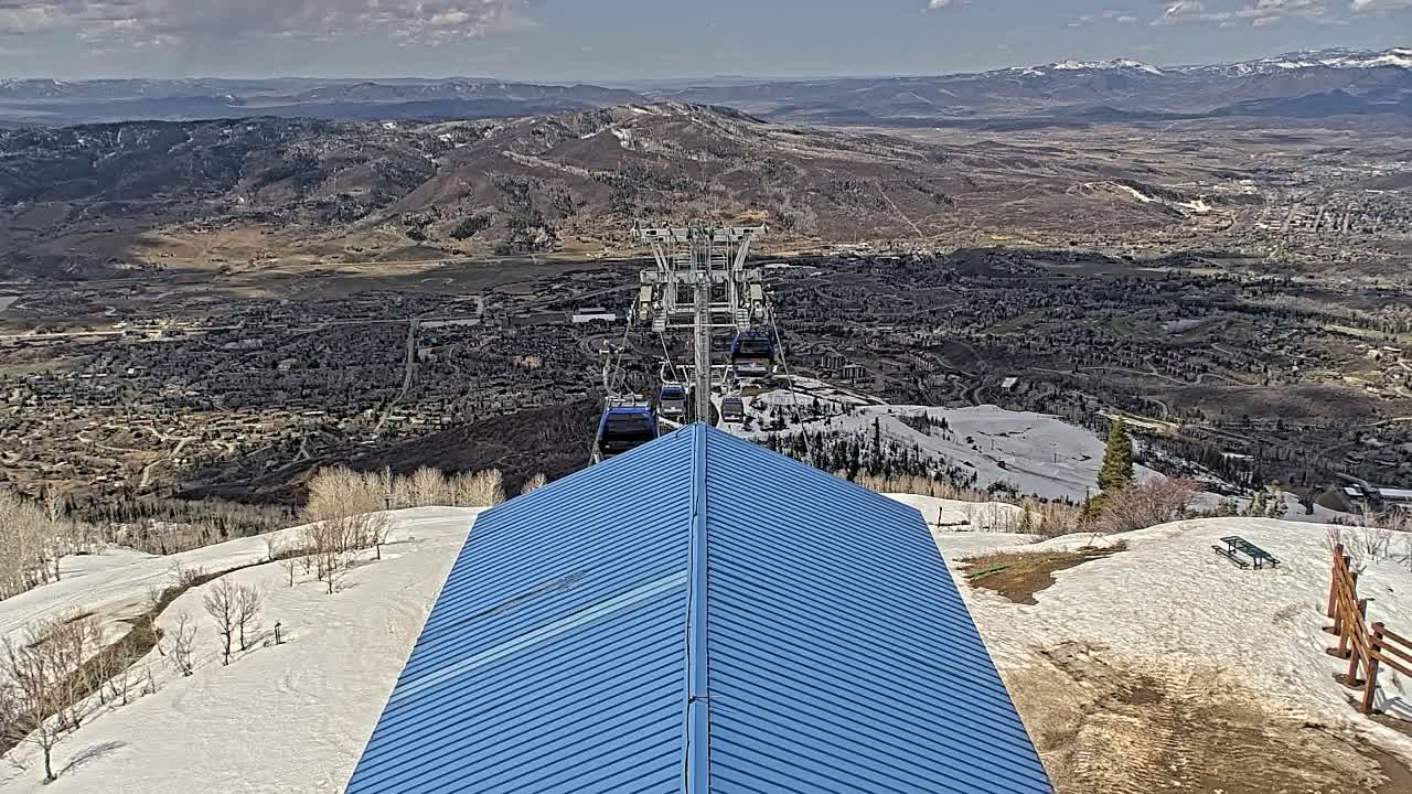 A blue metal roof is centered in the foreground, overlooking a snowy mountain slope, a ski lift carrying several gondolas, and a sprawling town nestled in a valley beneath a mostly clear sky.