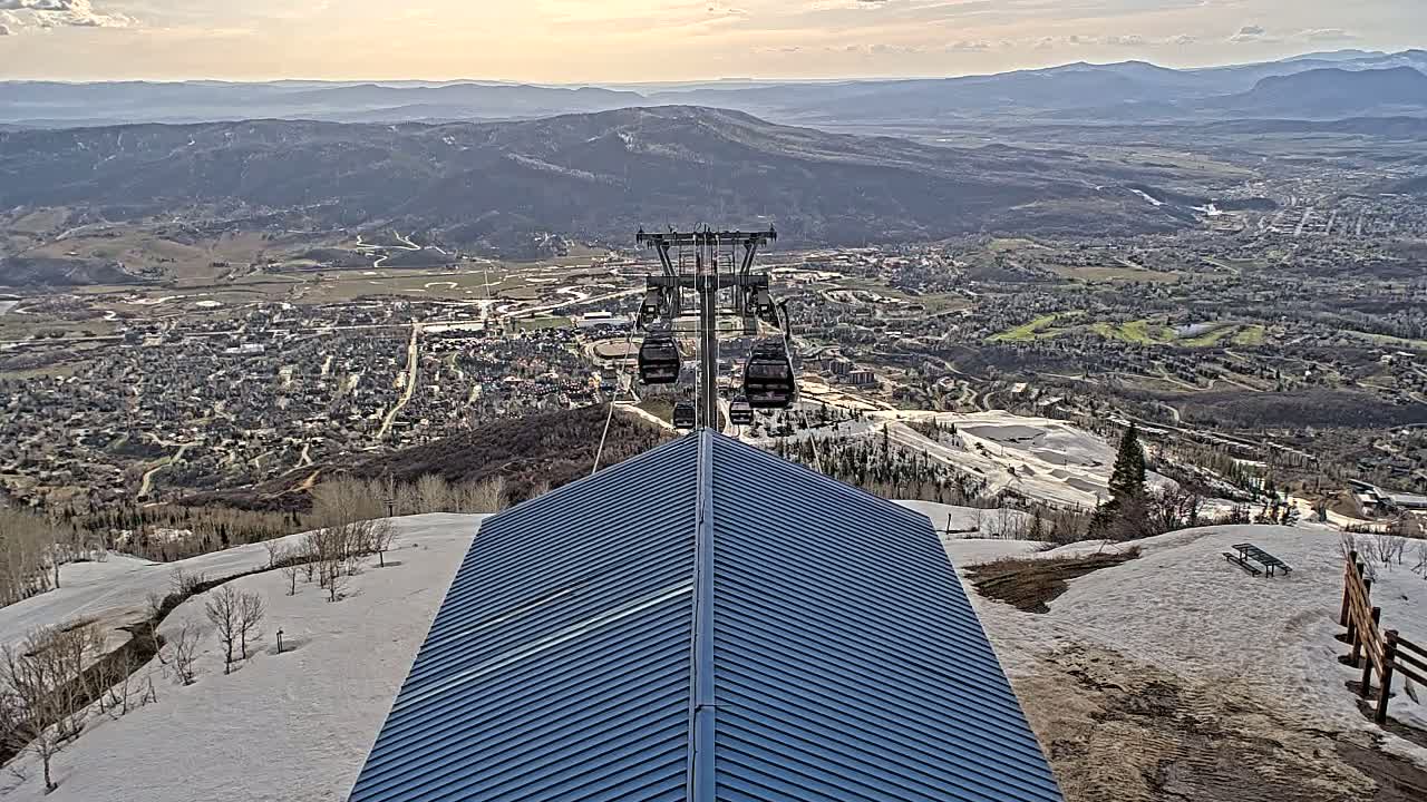A ski lift ascends from a blue-roofed building toward a valley town under partly cloudy skies, with patches of snow still visible on the mountain.