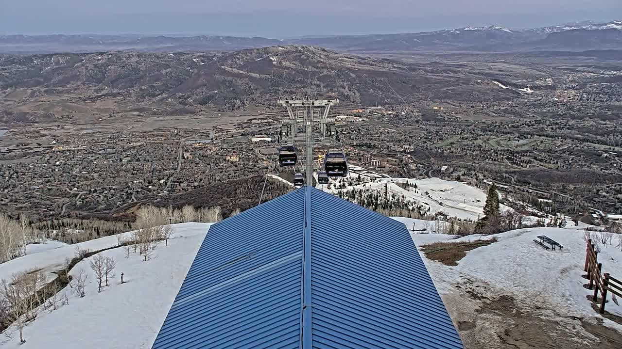 A blue-roofed building sits atop a snowy mountain overlooking a valley town under a mostly clear sky.