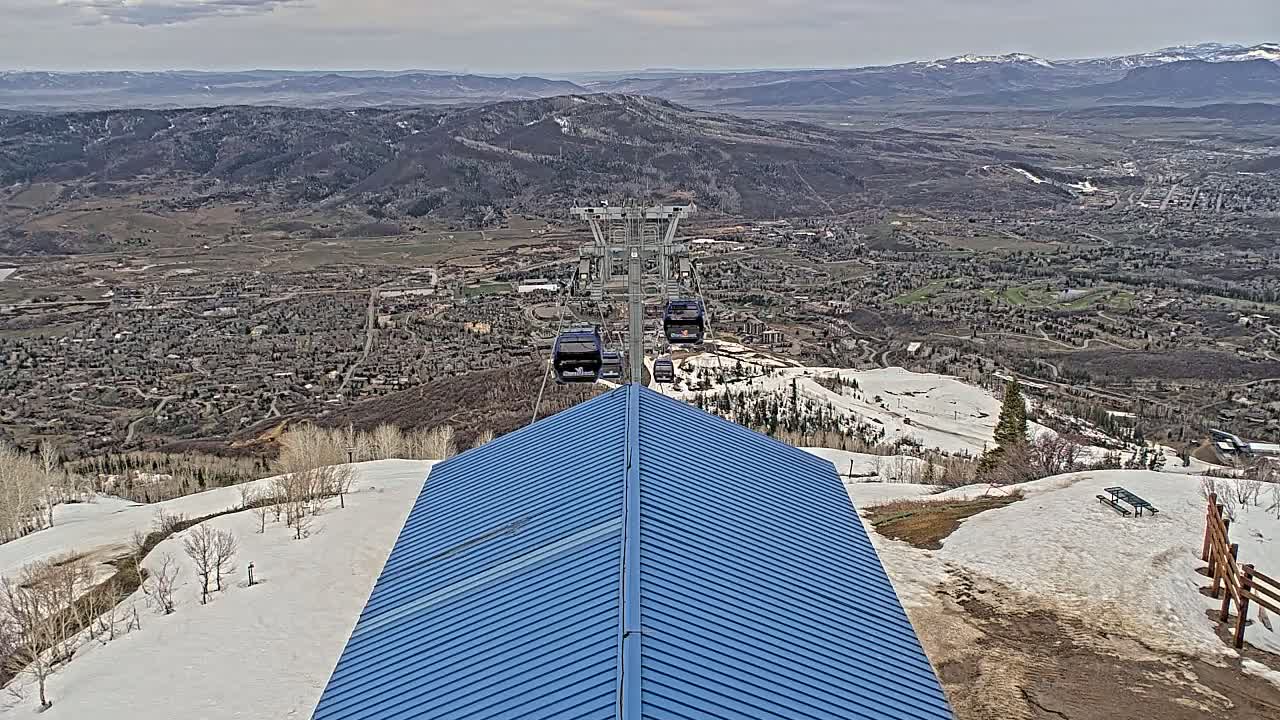 A blue-roofed building sits atop a snowy mountain overlooking a valley town under a partly cloudy sky.