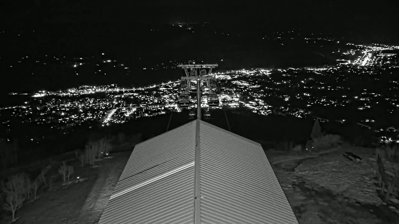 A nighttime view from atop a ridged roof shows a chairlift ascending toward a distant, brightly lit town under a dark sky.