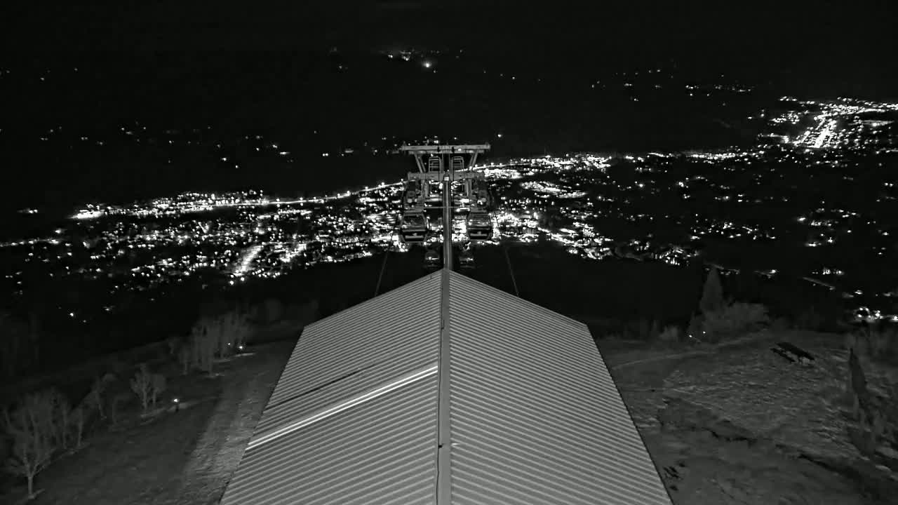 At night, a cable car ascends over a snow-covered landscape overlooking a brightly lit city.