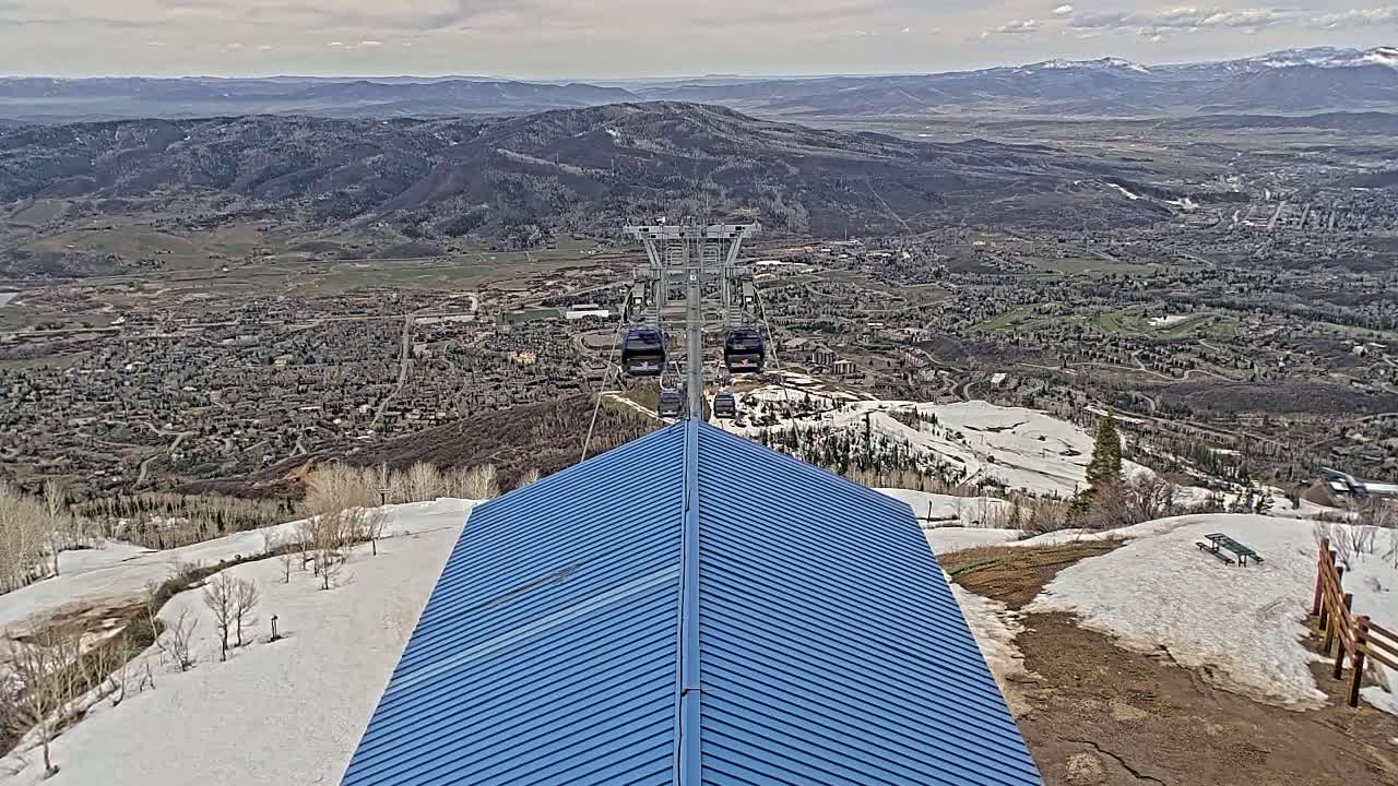 A blue-roofed structure overlooks a mountain town and ski resort under partly cloudy skies, with a gondola lift visible in the distance.