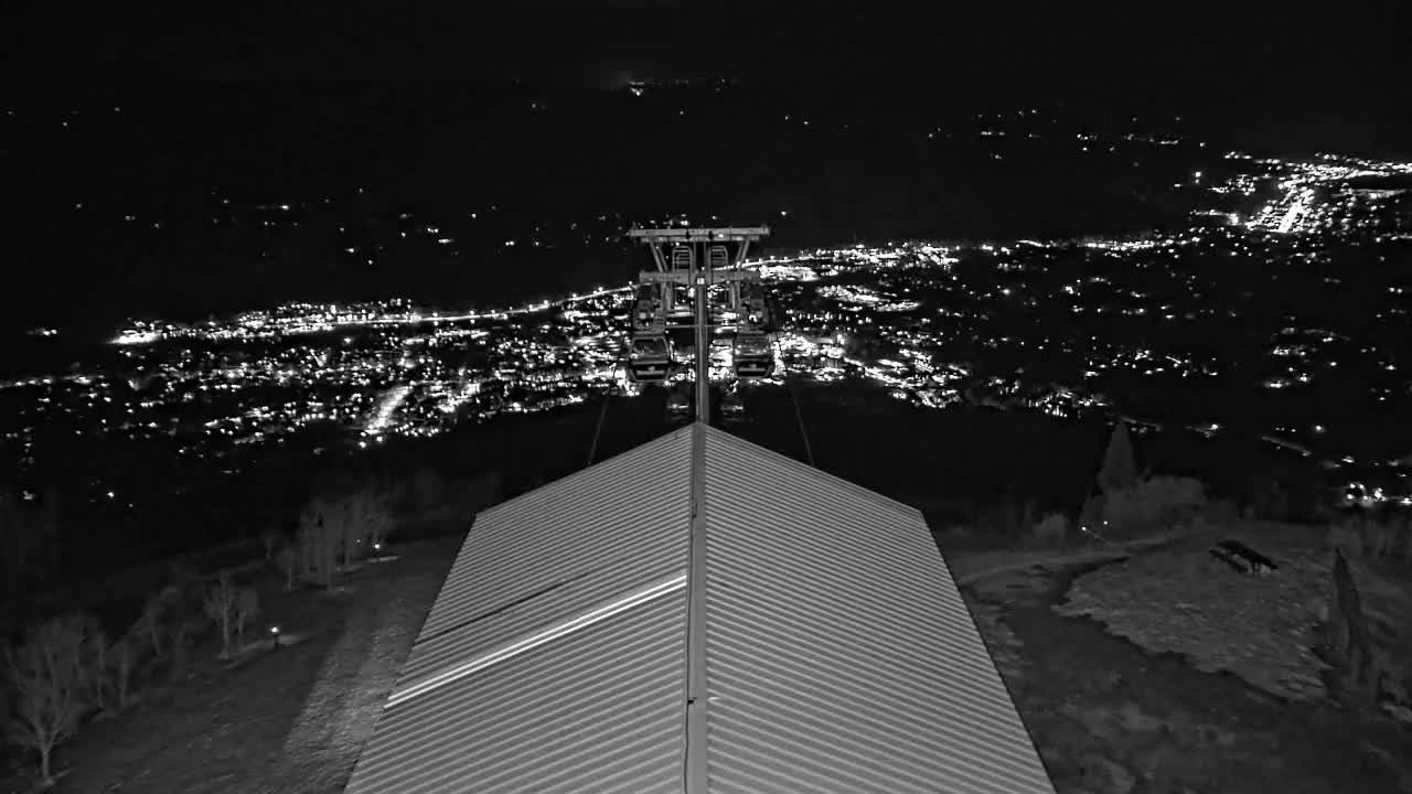 A nighttime view from atop a ridged roof shows a cable car ascending towards a distant, brightly lit town.