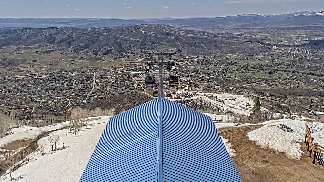 A blue roofed structure overlooks a valley town with a chairlift in the distance, under clear skies with patches of snow on the ground.