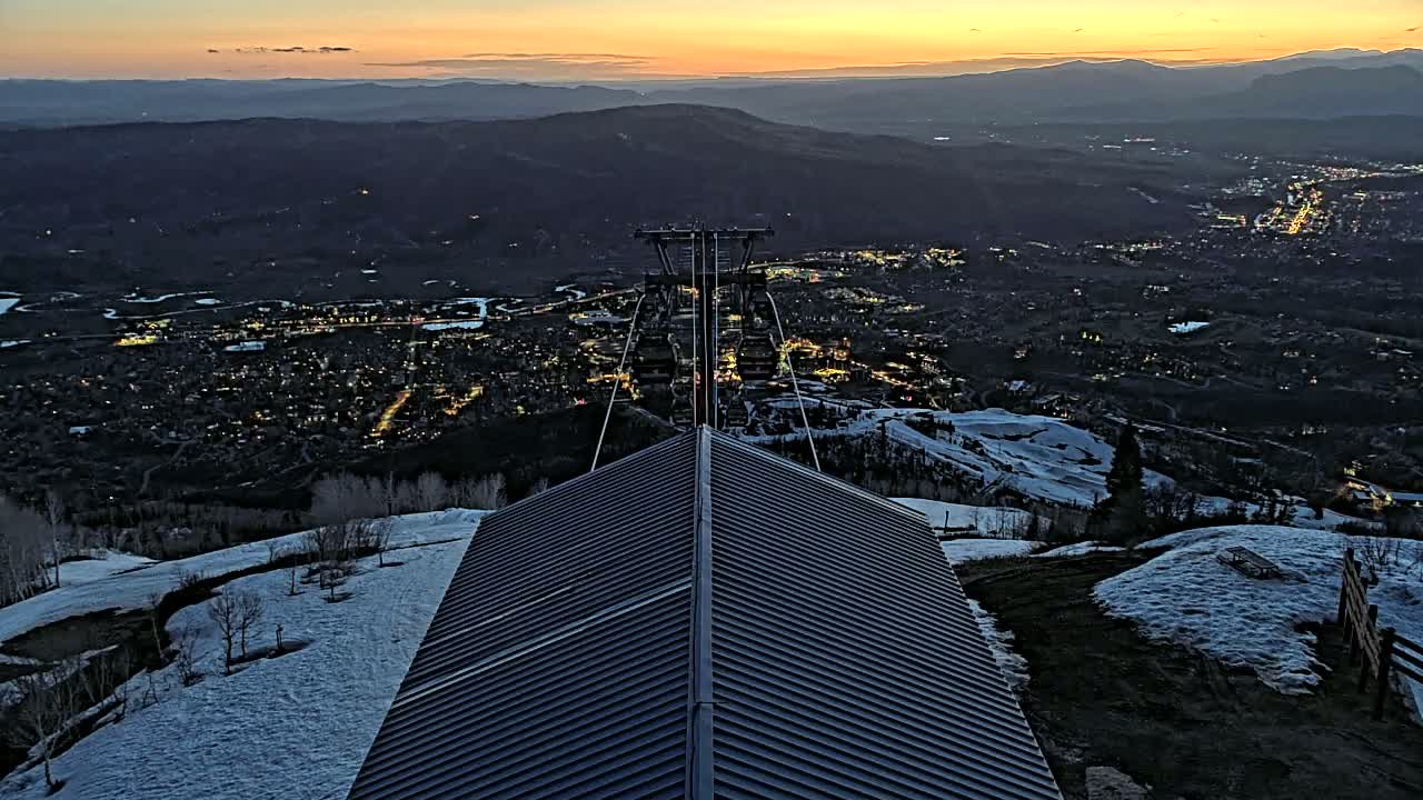 A dark gray, peaked roof sits atop a snowy mountain at sunset overlooking a valley town illuminated by lights.