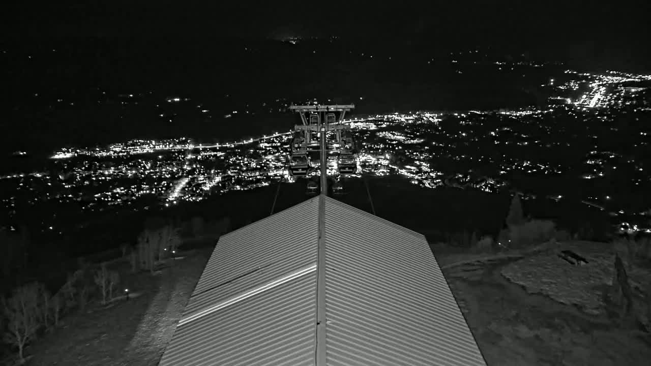 A nighttime view from atop a building shows a chairlift ascending toward a distant, brightly lit town, with a light dusting of snow on the ground.