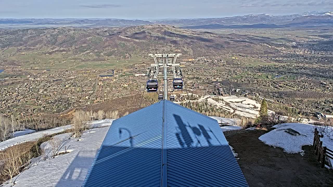 A mountaintop view on a sunny day shows a ski resort town below with a chairlift ascending, partially snow-covered terrain, and a dark-blue peaked roof in the foreground.