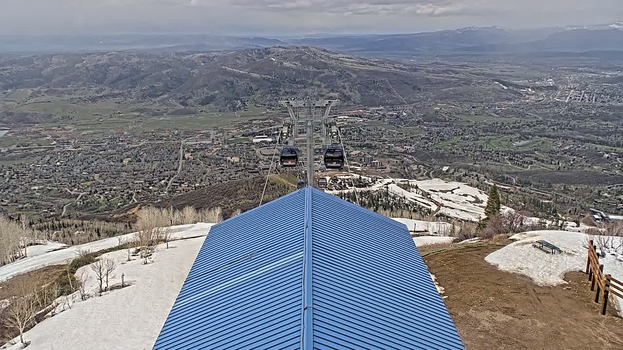 A blue-roofed structure overlooks a valley town with a chairlift ascending a mountain in the background, under overcast skies with patches of snow on the ground.