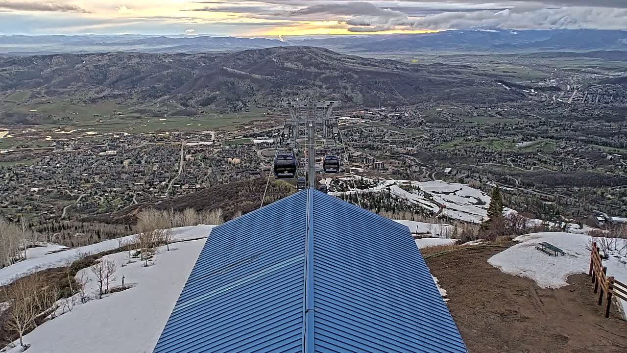 A blue metal roofed structure overlooks a valley town with a chairlift and patches of snow under partly cloudy skies.