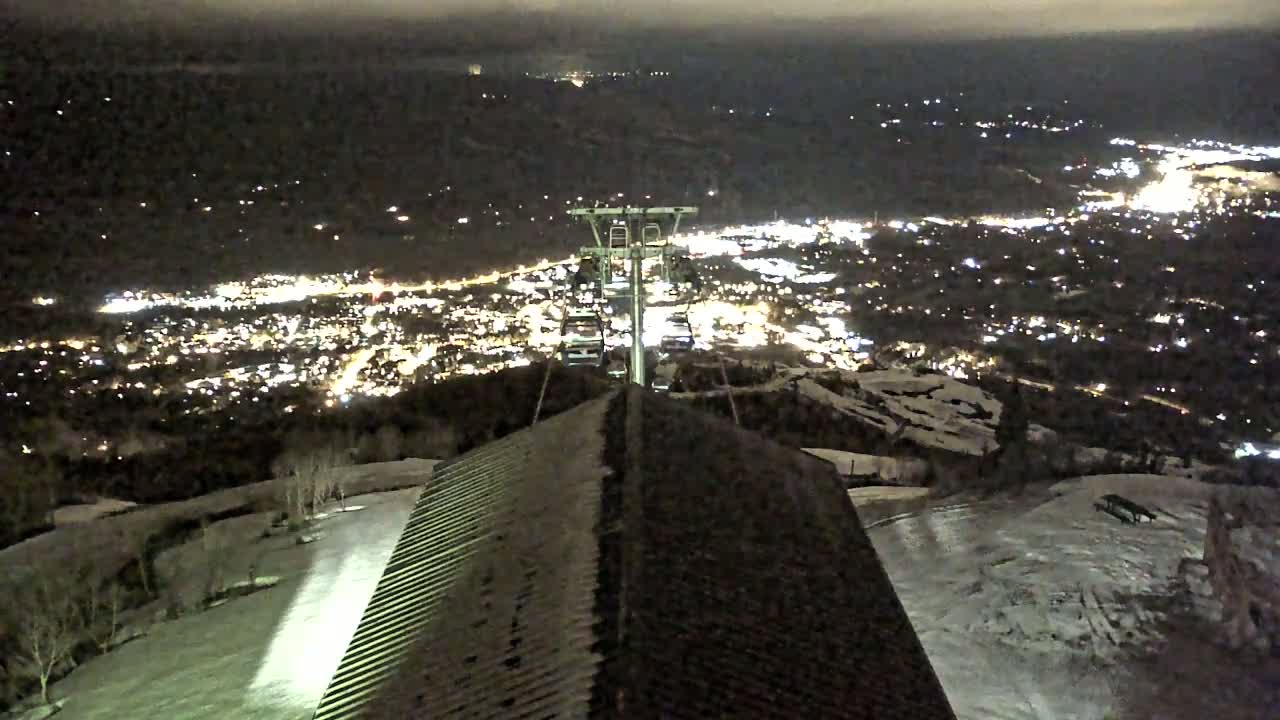 A nighttime view from a ski lift shows a snow-covered mountain slope, a partially snow-covered building's roof, and a brightly lit town in the valley below.