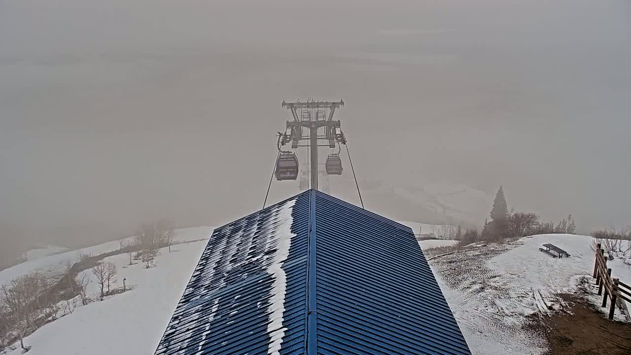 A chairlift ascends through a foggy, snowy mountain landscape towards a building with a dark blue, snow-dusted roof.