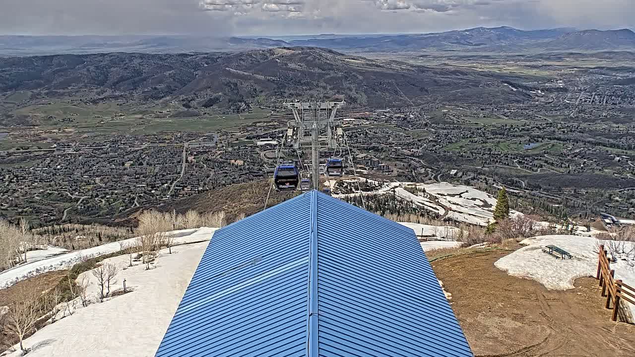A blue roof overlooks a ski resort town under a partly cloudy sky, with a chairlift visible in the distance and patches of snow remaining on the mountainside.