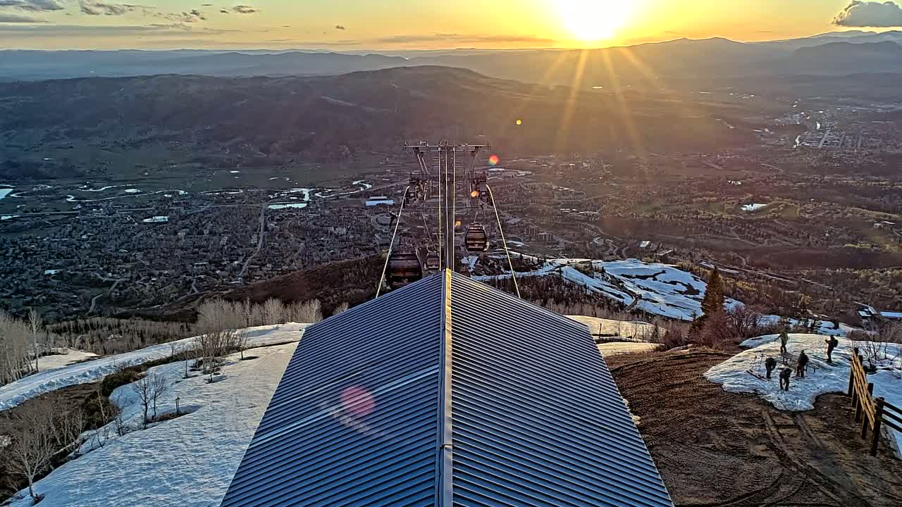 A mountaintop view at sunset shows a ski lift ascending toward the sun over a town nestled in a valley with patches of snow still visible on the mountainside.