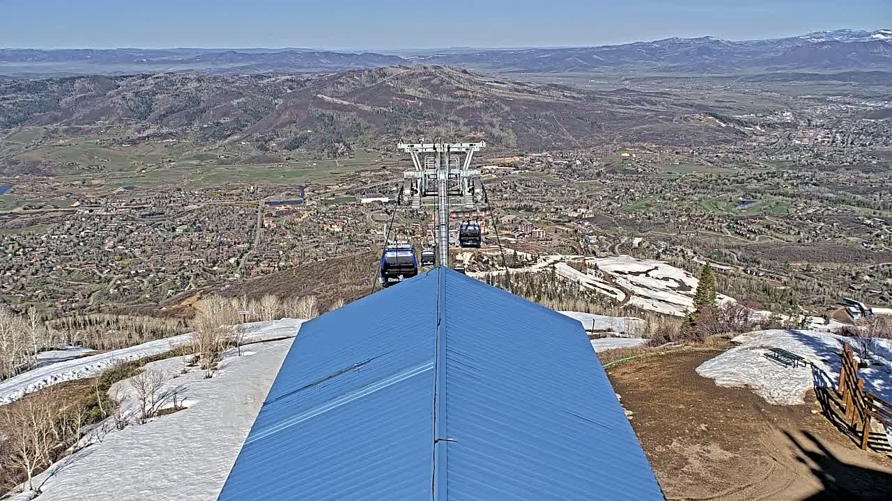 A blue roof overlooks a ski resort town nestled in a valley on a clear, sunny day, with a chairlift carrying several gondolas visible in the distance.