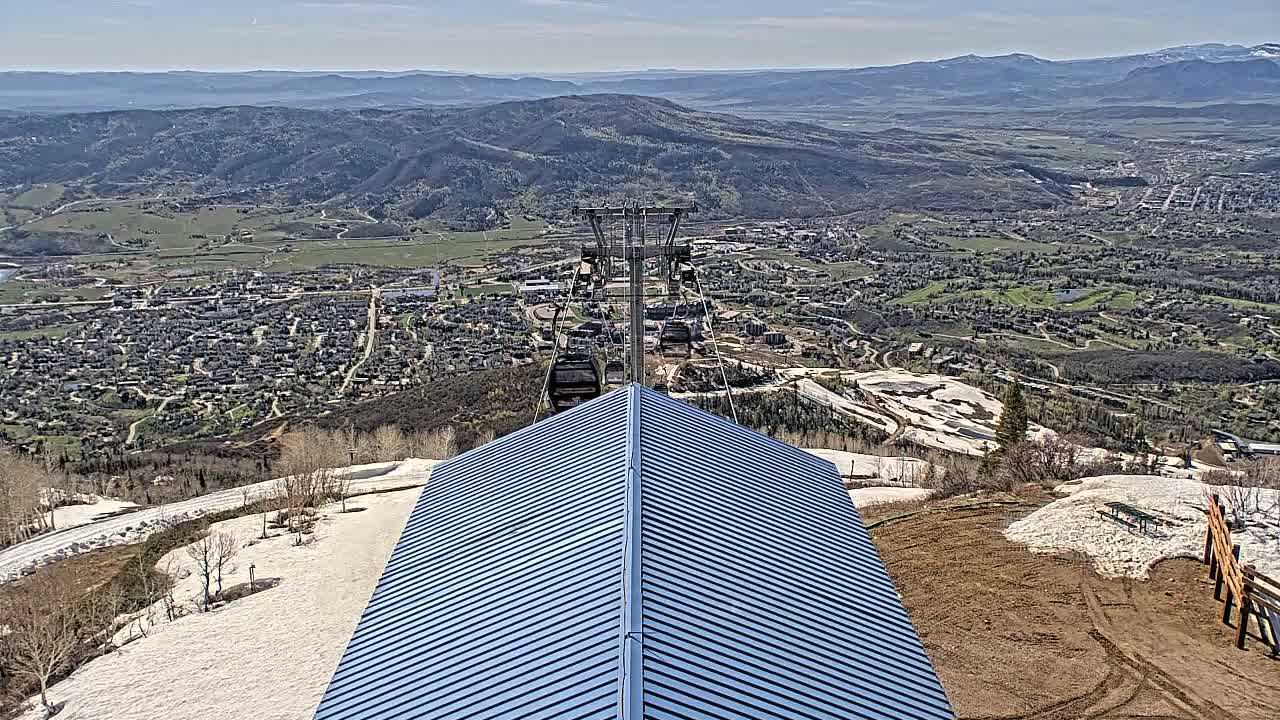 A blue metal roof sits atop a mountain overlooking a valley town, with snow patches on the ground and a chairlift in the background under a mostly clear sky.