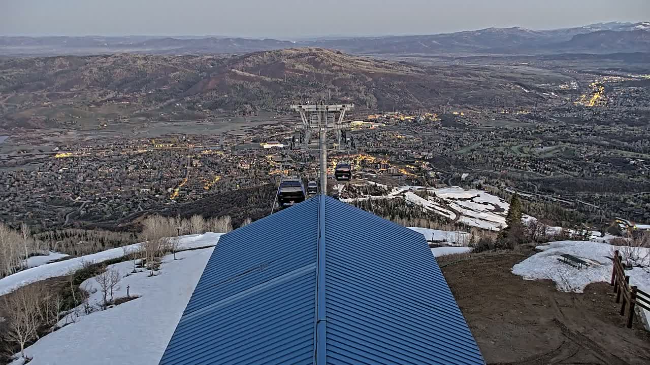 A blue-roofed structure overlooks a valley town with lights illuminated at dusk, showing patches of snow on the surrounding mountainside and several ski lift gondolas descending.