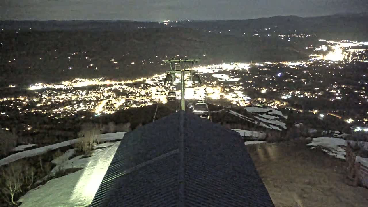 A nighttime view from a ski lift shows a city illuminated below, with patches of snow visible on the surrounding slopes.