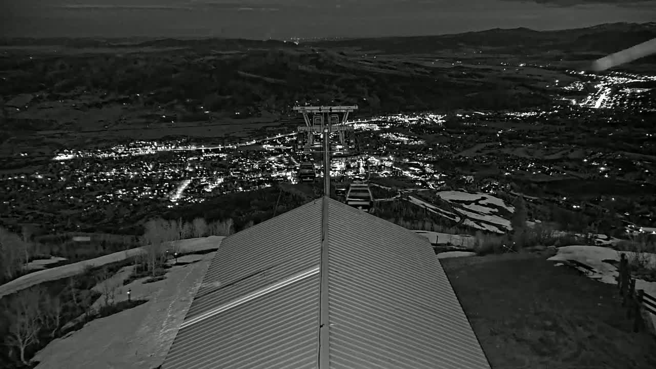 A nighttime view from a snow-dusted mountaintop shows a metal roof, a ski lift, and a valley town illuminated below.