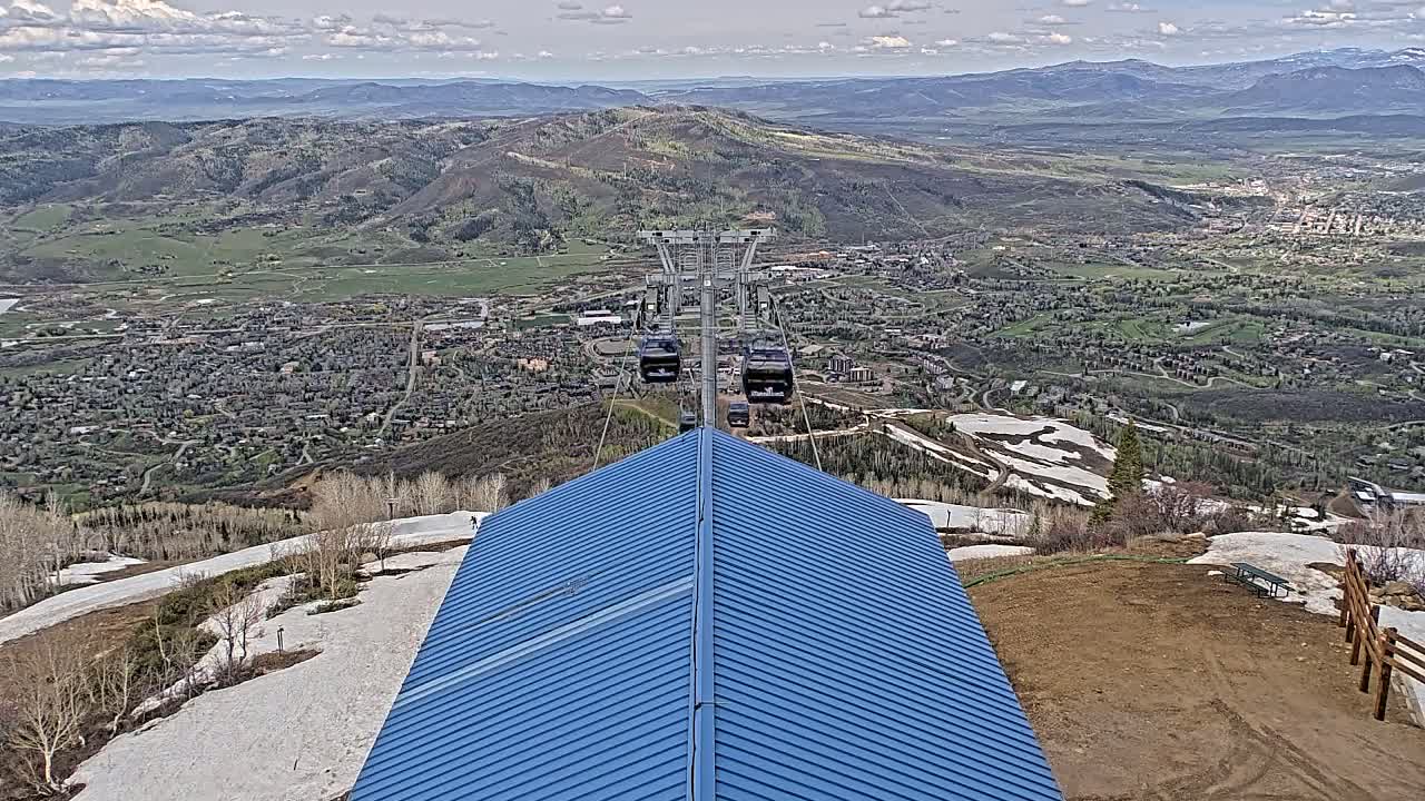 A blue-roofed building sits atop a mountain overlooking a valley town with patches of snow on the ground under partly cloudy skies.